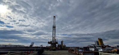 Oil drilling rig on a worksite with stacked pipes and trucks under a cloudy sky.