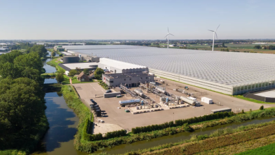 Aerial view of an agricultural processing complex with large greenhouses, processing buildings, and wind turbines in a rural landscape beside canals.