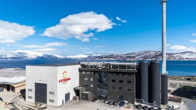 Industrial campus by a blue fjord with snow-covered mountains in the distance; white and dark gray buildings with large silos and a tall smokestack on the right side.