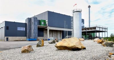 Industrial facility with grey and blue buildings, a tall silver silo and smokestack, and a rocky, gravel foreground.