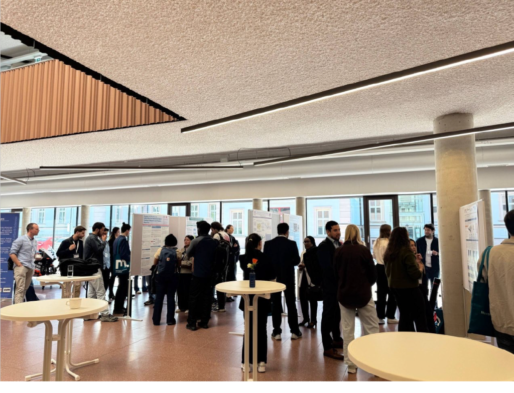 Group of people at a poster presentation in a bright, open hall with tall windows and round high tables.