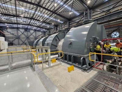 Steam-turbine | JeotermalHaberler - Jeotermal Haber Industrial power plant scene with large gray turbine-like machines lined along a warehouse, safety yellow rails, and overhead lighting.