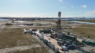 Aerial view of a coastal industrial site with a tall red-and-white communications tower, containers, and vehicles around a fenced work area, city skyline in the distance.