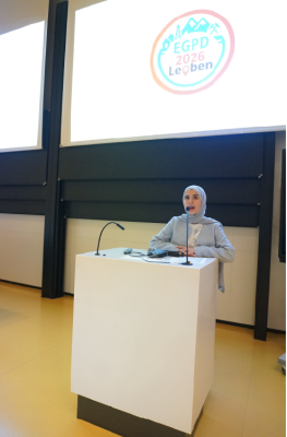 Female speaker wearing a hijab stands at a white podium with microphones in a lecture hall, presenting to an audience.