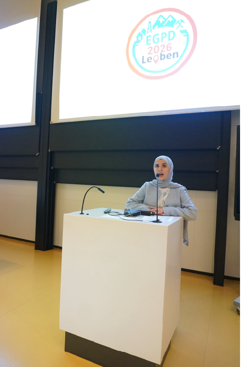 Female speaker wearing a hijab stands at a white podium with microphones in a lecture hall, presenting to an audience.