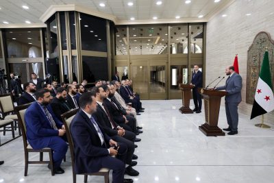Formal event or press conference with two podiums and a speaker addressing a seated audience in a marble-floored room, flags visible to the right
