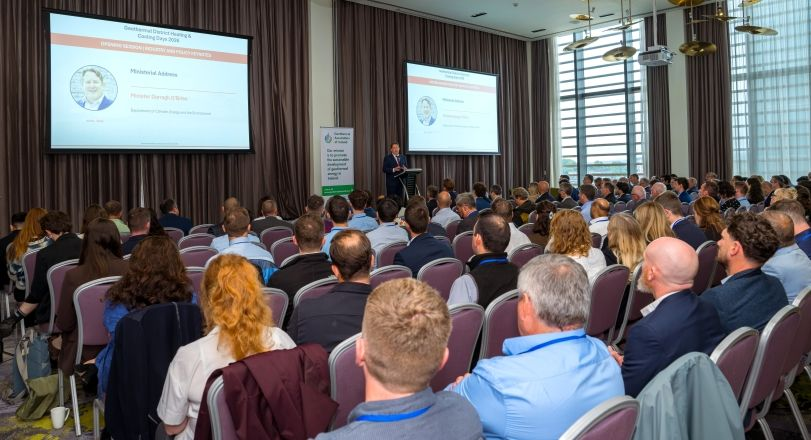 Darragh-Dublin | JeotermalHaberler - Jeotermal Haber Presenter giving a talk at a conference podium with two projection screens displaying slides, facing a large audience in a ballroom-style conference room.