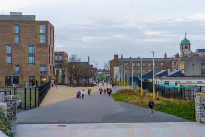 Urban walkway between modern brick buildings, with people strolling along a paved path and a distant clock tower on the right.