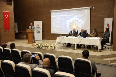 Speaker at a lectern delivering a presentation in a conference hall, with a large projection screen and panelists at a long table on stage.