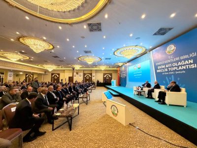 83857-59405-20261019132424 | JeotermalHaberler - Jeotermal Haber Panel discussion in a grand conference hall with a blue stage backdrop and seated speakers, audience in formal attire nearby.