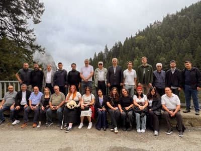 Sumela | JeotermalHaberler - Jeotermal Haber Large group of diverse adults posing for a group photo outdoors, with a forested mountainside and cloudy sky behind them.